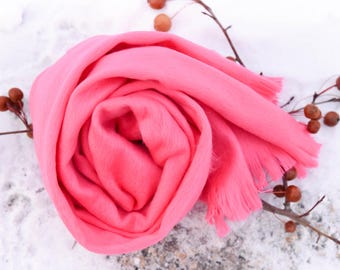 May include: A vibrant pink scarf, artfully arranged in a spiral, with fringed edges. The soft fabric is set against a snowy backdrop, with sprigs of berries adding a touch of natural detail. A cozy accessory.