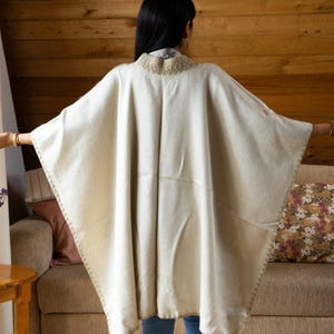 May include: Cream-colored poncho with a decorative collar and stitched edges. The poncho is made of a soft, woven material and is worn over a light-colored shirt and blue jeans. The background features a wooden wall and a sofa.