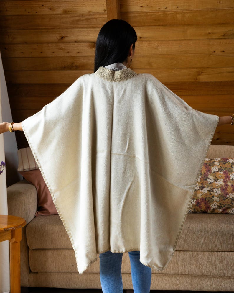 May include: Cream-colored poncho with a decorative collar and stitched edges. The poncho is made of a soft, woven material and is worn over a light-colored shirt and blue jeans. The background features a wooden wall and a sofa.
