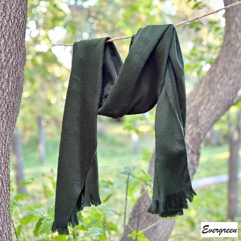 May include: A dark green scarf with fringed edges hangs from a washing line. The scarf is made of a soft material and is draped in a relaxed style. The word "Evergreen" is in the bottom right corner.