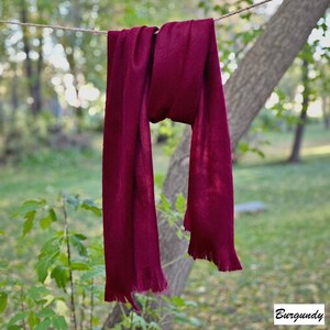 May include: A burgundy scarf hangs from a washing line against a blurred green backdrop. The scarf is made of a soft material and has a fringed edge. The word "Burgundy" is in the bottom right corner.