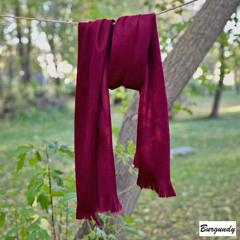 May include: A burgundy scarf hangs from a washing line against a blurred green backdrop. The scarf is made of a soft material and has a fringed edge. The word "Burgundy" is in the bottom right corner.