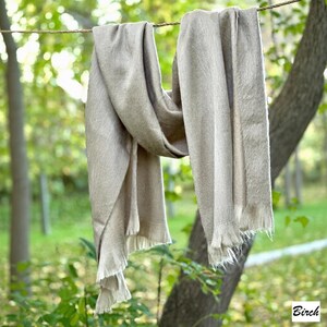 May include: A beige scarf with fringed edges hangs from a rope outdoors. The scarf is draped over the rope, showing its texture and design. The background features a blurred view of trees and foliage. The word "Birch" is in the bottom right corner.