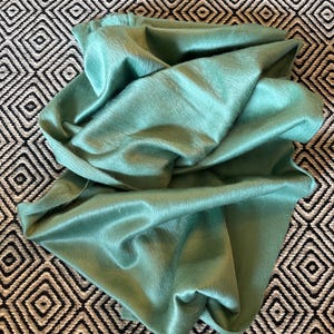 May include: A folded, seafoam green textile with a soft, furry texture. The fabric is draped over a black and white geometric patterned surface. The textile has a subtle sheen, suggesting a luxurious material.