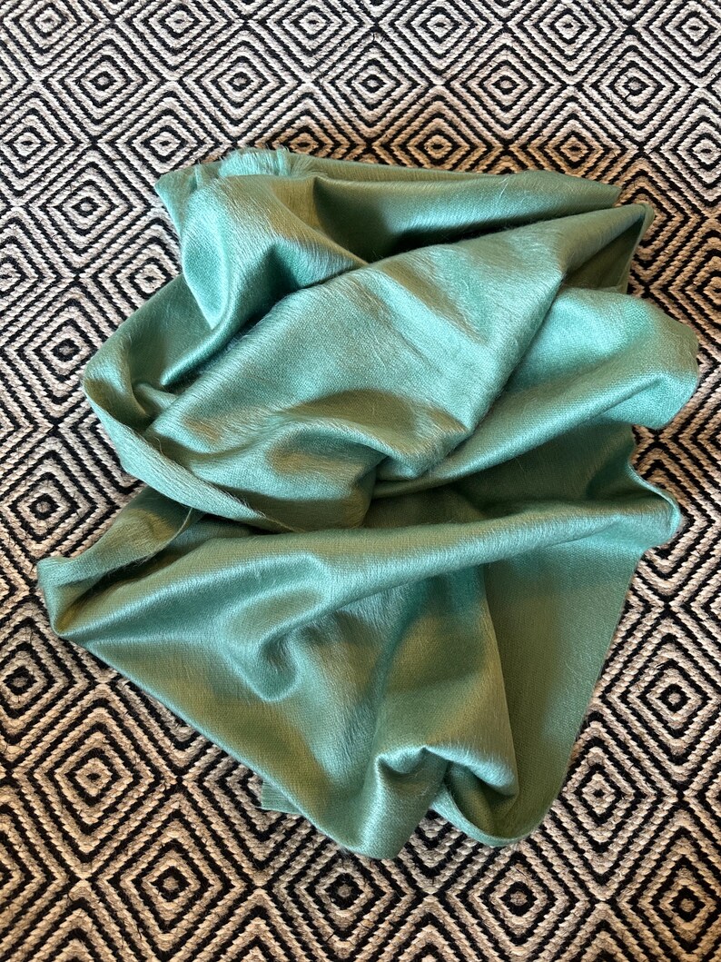 May include: A folded, seafoam green textile with a soft, furry texture. The fabric is draped over a black and white geometric patterned surface. The textile has a subtle sheen, suggesting a luxurious material.