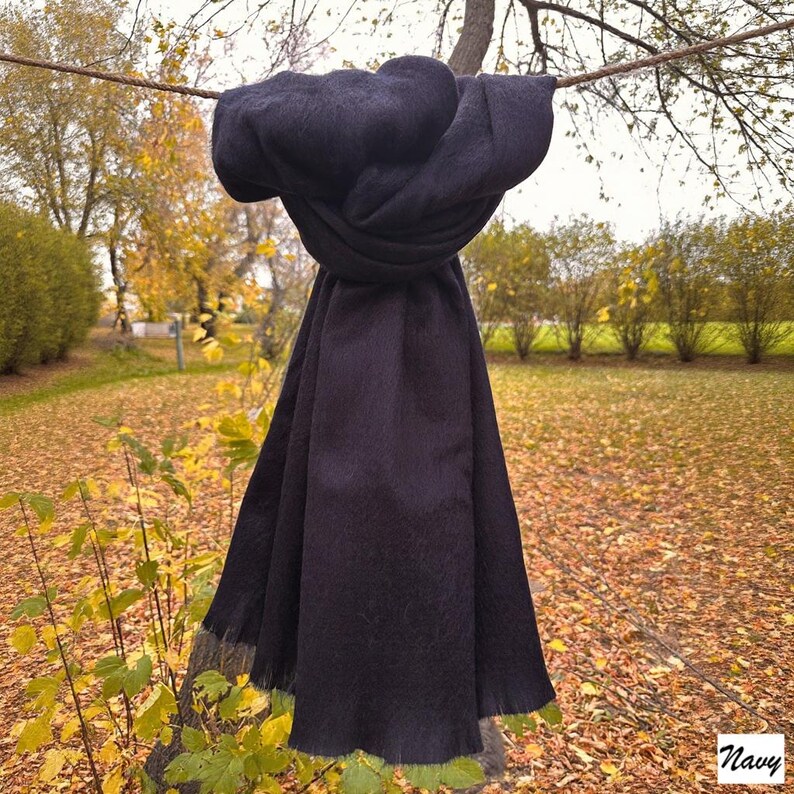 May include: A navy blue scarf, knotted and draped over a branch, displays its soft texture and fringed edge. The word "Navy" is in the bottom right corner. The background shows an autumnal scene.