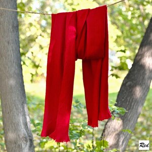 May include: A bright red scarf hangs on a washing line between two trees. The scarf is made of a soft material and has a fringed edge. The background is blurred, with green foliage and sunlight.