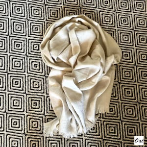 May include: A cream-coloured scarf with a fringed edge is draped over a patterned surface. The scarf appears soft and is made of a lightweight material. The background features a repeating geometric design in black and white.