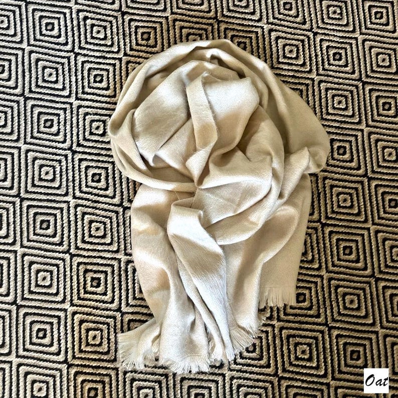 May include: A cream-coloured scarf with a fringed edge is draped over a patterned surface. The scarf appears soft and is made of a lightweight material. The background features a repeating geometric design in black and white.