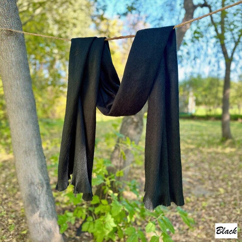 May include: A black scarf hangs from a washing line, with the word "Black" in a white box in the lower right corner. The scarf is made of a soft material and has a fringed edge. The background is a blurred view of trees and greenery.