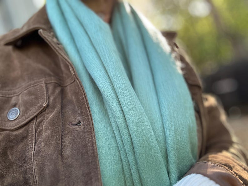 May include: A close-up of a brown suede jacket with a light blue scarf. The jacket has a button closure and a pocket on the chest. The scarf is draped around the neck and has a soft, textured appearance.