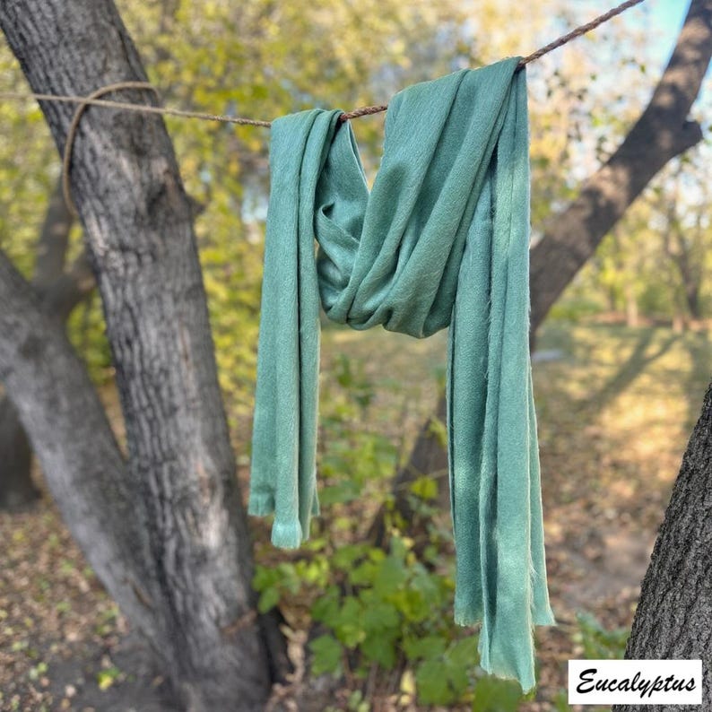 May include: A soft, eucalyptus-coloured scarf is draped over a rope, hanging between two trees. The scarf has a long, rectangular shape with a slightly frayed edge. The word "Eucalyptus" is printed in a white box in the bottom right corner.