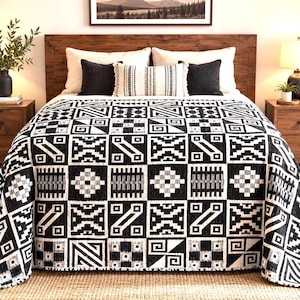 May include: A black and white bedspread featuring a repeating geometric pattern of squares, triangles, and lines. The textile is draped over a bed with two solid black square pillows and one rectangular white pillow with black vertical stripes.