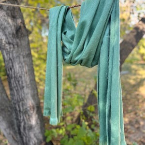May include: A soft, sage green scarf is draped over a rope, hanging in front of a blurred background of trees and foliage. The scarf's fabric appears lightweight and flowing, with a slightly textured surface. The scarf is a solid color.