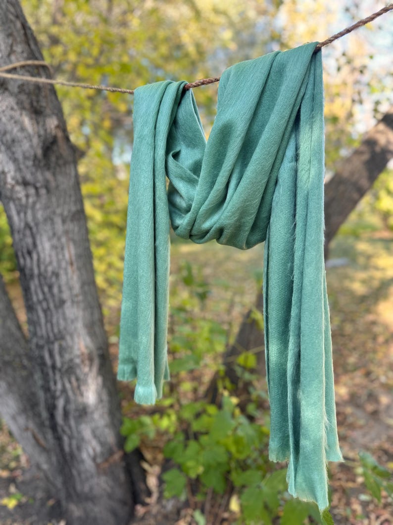 May include: A soft, sage green scarf is draped over a rope, hanging in front of a blurred background of trees and foliage. The scarf's fabric appears lightweight and flowing, with a slightly textured surface. The scarf is a solid color.