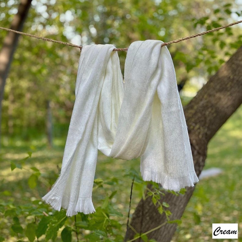 May include: A cream-coloured scarf with fringed edges, hanging from a rope against a blurred green backdrop. The scarf is made of a soft, lightweight material. The word "Cream" is in the bottom right corner.
