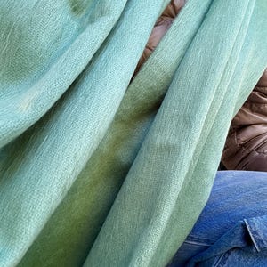 May include: A close-up shot of a soft, light green scarf. The scarf is draped in folds, revealing a textured weave. The color transitions from a lighter shade to a deeper green. The scarf appears to be made of a lightweight, possibly natural, material.