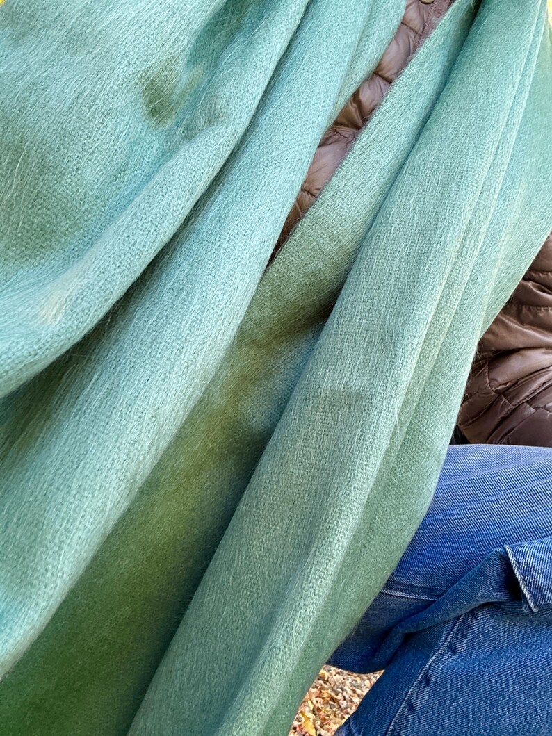 May include: A close-up shot of a soft, light green scarf. The scarf is draped in folds, revealing a textured weave. The color transitions from a lighter shade to a deeper green. The scarf appears to be made of a lightweight, possibly natural, material.