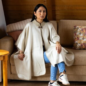 May include: A cream-colored cape with a decorative collar and two wooden buttons. The cape has a rounded hem and is worn over blue jeans. The person is sitting on a brown sofa in a room with wood paneling.