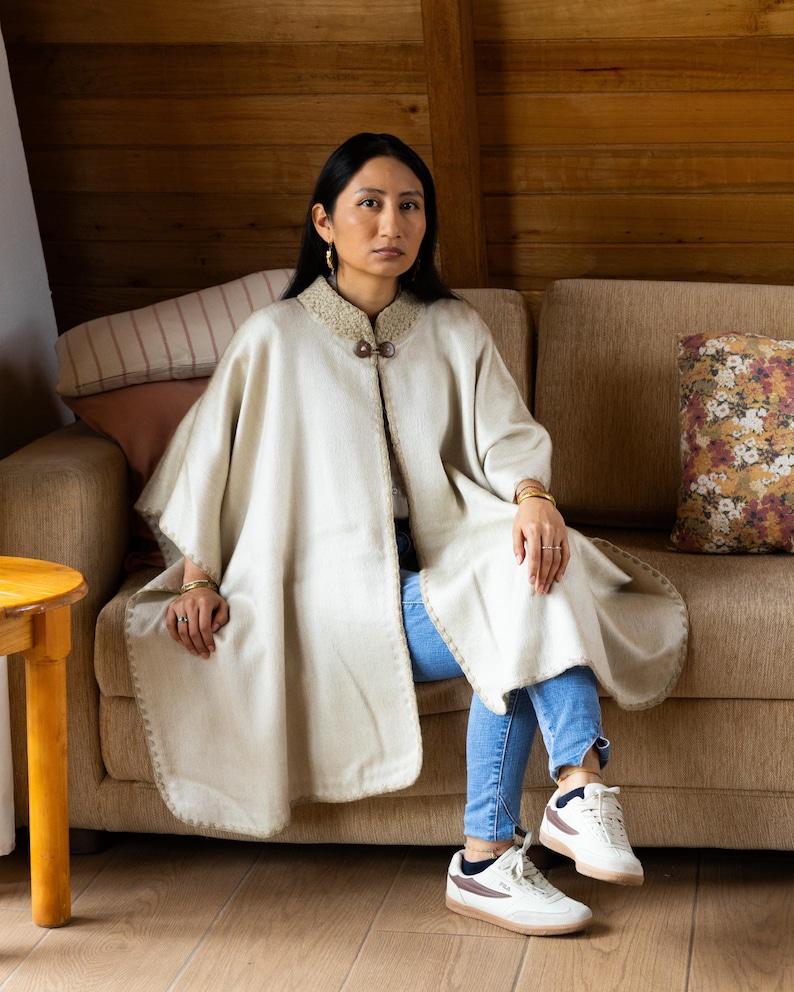 May include: A cream-colored cape with a decorative collar and two wooden buttons. The cape has a rounded hem and is worn over blue jeans. The person is sitting on a brown sofa in a room with wood paneling.