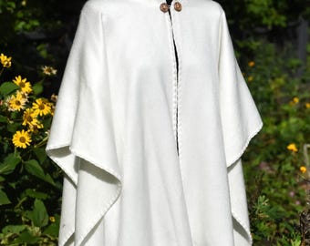 Op de afbeelding: Een witte cape met een hoge kraag en twee houten knopen. De cape is gemaakt van een zachte, getextureerde stof en heeft een vloeiend ontwerp. De achtergrond toont groen gebladerte en gele bloemen.