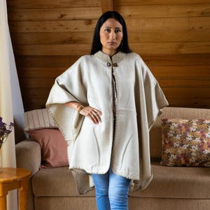 May include: A cream-colored poncho with a decorative collar and wooden buttons. The poncho has a stitched border and is worn over a white top and blue jeans. The background features a wooden wall and a sofa with decorative pillows.