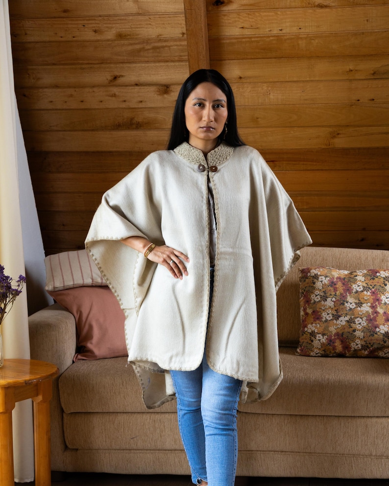 May include: A cream-colored poncho with a decorative collar and wooden buttons. The poncho has a stitched border and is worn over a white top and blue jeans. The background features a wooden wall and a sofa with decorative pillows.