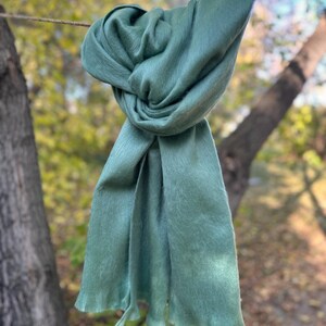 May include: A sage green scarf is knotted and draped, hanging from a thin rope. The scarf's fabric appears soft and flowing, with a slightly textured surface. The background features blurred trees and foliage, suggesting an outdoor setting.