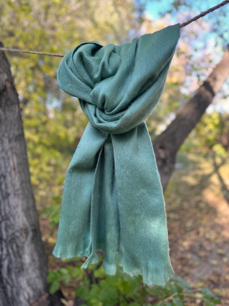 May include: A sage green scarf is knotted and draped, hanging from a thin rope. The scarf's fabric appears soft and flowing, with a slightly textured surface. The background features blurred trees and foliage, suggesting an outdoor setting.