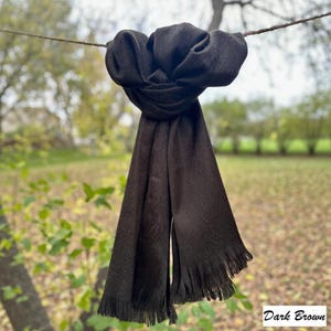 May include: A dark brown scarf with fringed edges hangs from a line against a blurred outdoor background. The scarf is knotted at the top, with the ends hanging down. The text "Dark Brown" is in the bottom right corner.