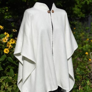 May include: A white cape with a high collar and two wooden buttons. The cape has a flowing design with a slightly textured surface. The garment is displayed on a black mannequin against a backdrop of green foliage and yellow flowers.