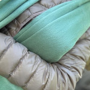 May include: A close-up shot of a teal-colored scarf draped over a tan puffer jacket. The scarf is soft and textured, with visible folds. The jacket has a quilted design and a shiny finish, creating a contrast with the scarf's texture. The overall aesthetic is cozy and warm.