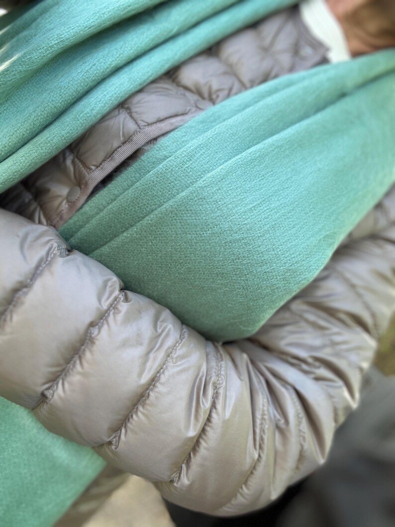 May include: A close-up shot of a teal-colored scarf draped over a tan puffer jacket. The scarf is soft and textured, with visible folds. The jacket has a quilted design and a shiny finish, creating a contrast with the scarf's texture. The overall aesthetic is cozy and warm.