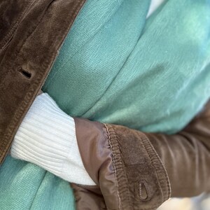 May include: A close-up of a brown suede jacket with a buttoned cuff, layered over a light blue scarf and a white sweater. The jacket has a classic design and a soft texture, creating a cozy and stylish look.