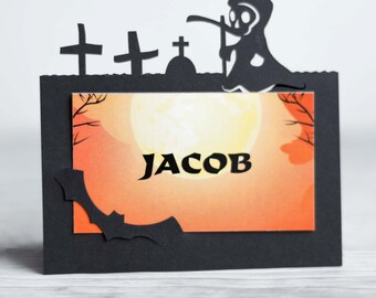 Könnte beinhalten: Ein schwarzer Halloween-Kartenhalter mit einem ausgeschnittenen Design mit Grabsteinen, einem Sensenmann und einer Fledermaus. Die Karte zeigt den Namen "JACOB" vor einem orange-gelben Hintergrund, der eine gruselige Atmosphäre erzeugt.