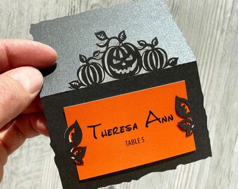 Könnte beinhalten: Eine schwarze und silberne Halloween-Tischkarte mit einem lasergeschnittenen Kürbisdesign. Der orangefarbene Einsatz trägt die Aufschrift "Theresa Ann" und "Table 5". Die Karte wird von einer Hand gehalten.