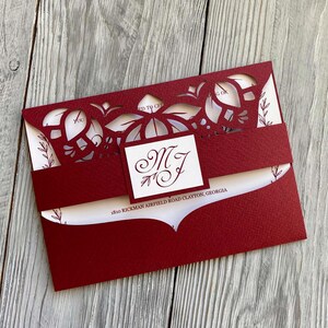 May include: Burgundy red wedding invitation with a white inner envelope and a laser cut design. The invitation features a monogram of the letters "M" and "J" in a cursive font. The address is printed on the bottom of the invitation: 1810 Rickman Airfield Road Clayton, Georgia.