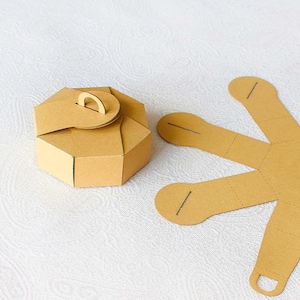 Może przedstawiać: Brązowe, ośmiokątne pudełko na prezent z uchwytem, obok płaskiego, gwiazdopodobnego kawałka kartonu. Pudełko jest złożone, podczas gdy drugi element jest niezłożony. Oba wykonane są z jasnobrązowego kartonu.