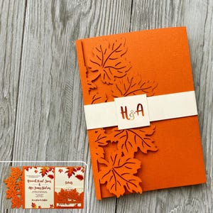 May include: Orange wedding invitation featuring a laser cut design of autumn leaves. The invitation has a white band with the initials "H&A" in gold lettering.