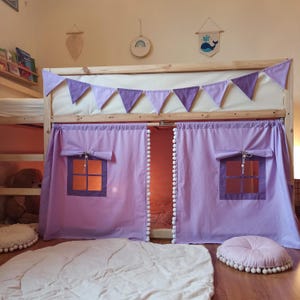 May include: A wooden loft bed frame featuring light purple fabric curtains with white pom-pom trim and two square windows. A row of purple and light purple triangular bunting hangs across the top rail. Two round, tufted light purple floor cushions.