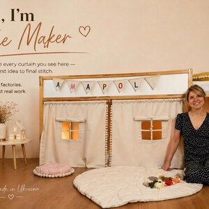 May include: A beige fabric playhouse curtain set attached to a wooden bed frame, featuring two windows with roll-up shades, a pom-pom trim, and a bunting banner spelling AMAPOLA. The curtain is made of solid light-colored fabric with stitched details.