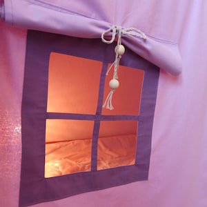 May include: A light purple fabric play tent featuring a square window with a dark purple frame. A rolled-up fabric shade is secured above the window with a white cord featuring two round wooden beads and a tassel at the end, creating a soft glow.