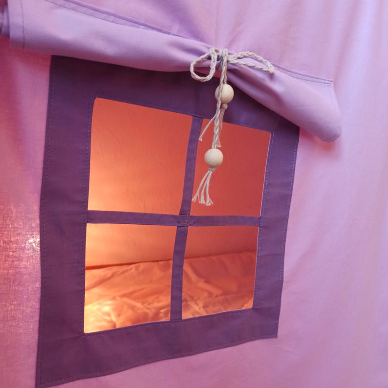 May include: A light purple fabric play tent featuring a square window with a dark purple frame. A rolled-up fabric shade is secured above the window with a white cord featuring two round wooden beads and a tassel at the end, creating a soft glow.