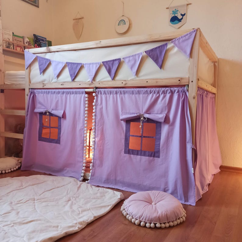 May include: A wooden loft bed frame featuring light purple fabric curtains with pom-pom trim, two roll-up windows with orange accents, and a matching purple triangular bunting banner draped across the top rail, accompanied by a round tufted floor cushion.