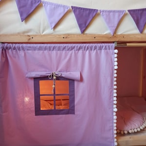May include: A light purple fabric curtain for a bed frame featuring a roll-up window with a dark purple frame, a tie-up closure with wooden beads, and a white pom-pom trim along the side. A string of purple triangular bunting hangs above.