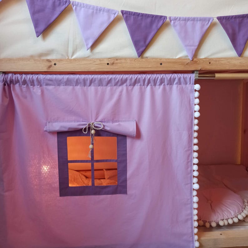 May include: A light purple fabric curtain for a bed frame featuring a roll-up window with a dark purple frame, a tie-up closure with wooden beads, and a white pom-pom trim along the side. A string of purple triangular bunting hangs above.
