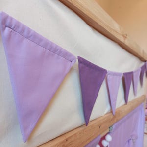 May include: A fabric bunting banner featuring a series of triangular flags in varying shades of purple, including light lavender and deep violet, suspended along a wooden frame. The pennants are stitched with a clean hem along the top edge.