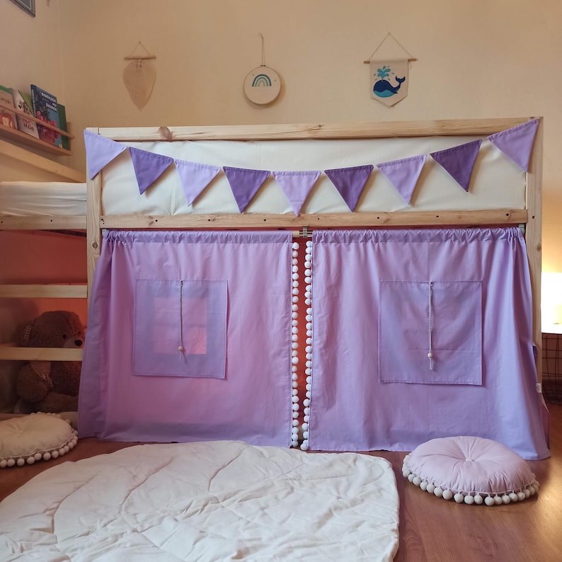 May include: A wooden loft bed frame featuring light purple fabric curtains with two square windows and white pom-pom trim. A matching light purple triangular bunting banner is attached to the top rail. Two round floor cushions with white pom-pom trim.