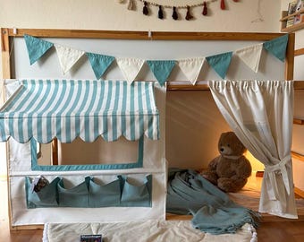 May include: A child's playhouse bed with a striped awning, teal and white pennant flags, and a round play mat with white pom-pom trim. A teddy bear sits inside the playhouse, with a blue blanket and a game box on the mat.
