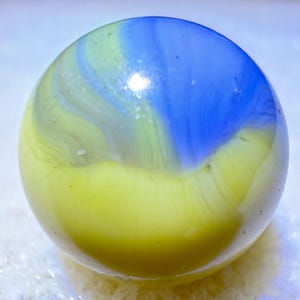 May include: A glass marble with a swirl of colours. The top half is blue, with a swirl of green and yellow on the side. The bottom half is yellow. The marble is round and smooth.